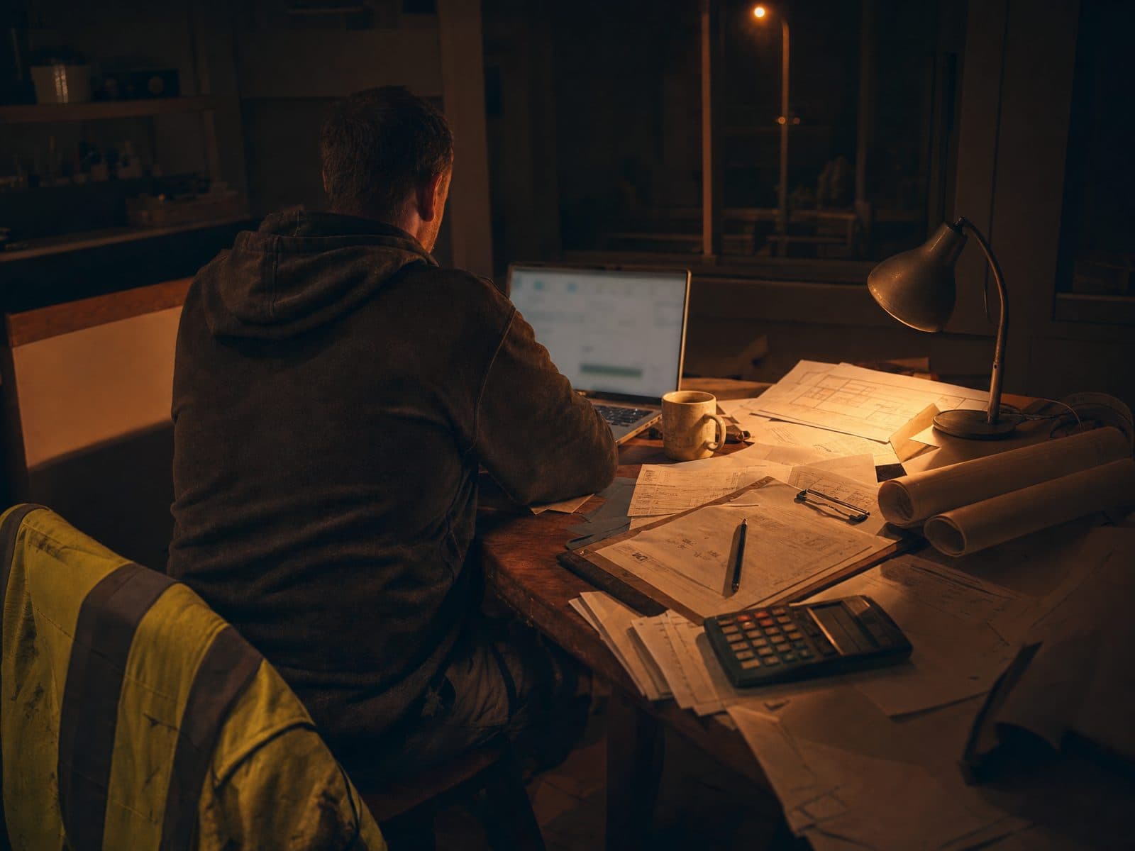 A builder from behind at the kitchen table late at night, laptop open, lamp on, surrounded by plans, timesheets and invoices.